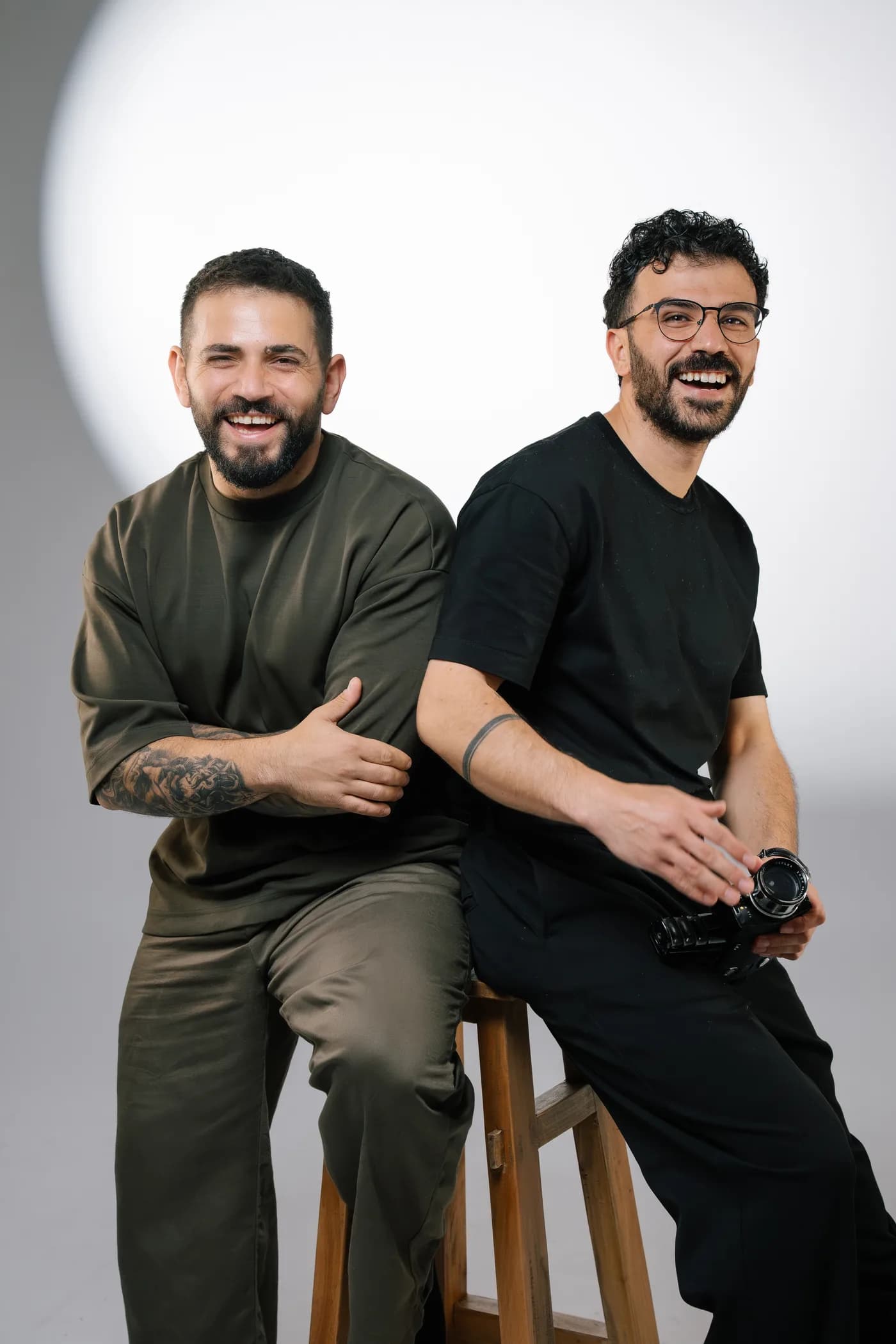 Sako und Adem von Studio Vielseitig, lachend Rücken an Rücken auf einem Hocker mit Kamera